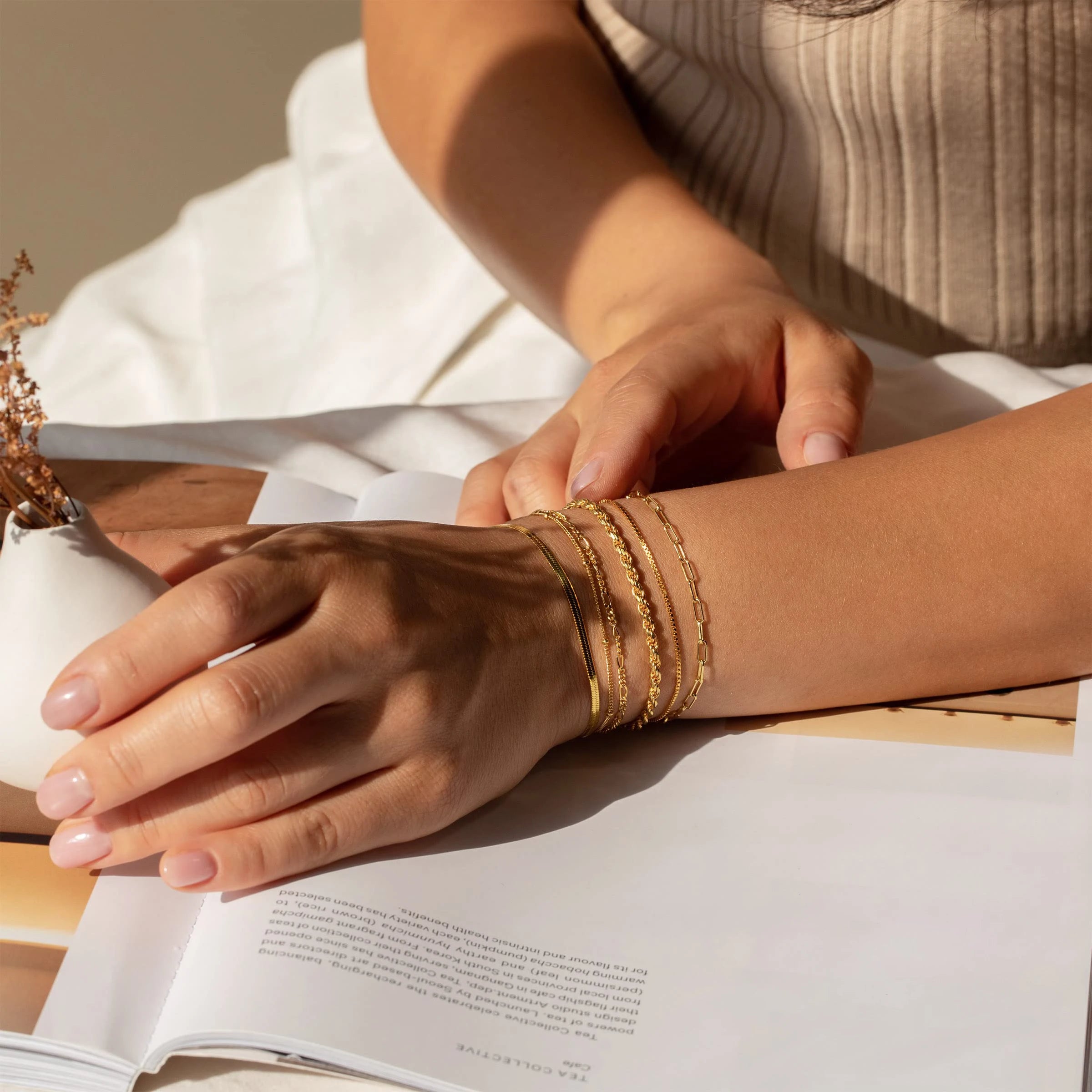 Close-up of a person's arm with gold bracelets on an open book, with a neutral background.