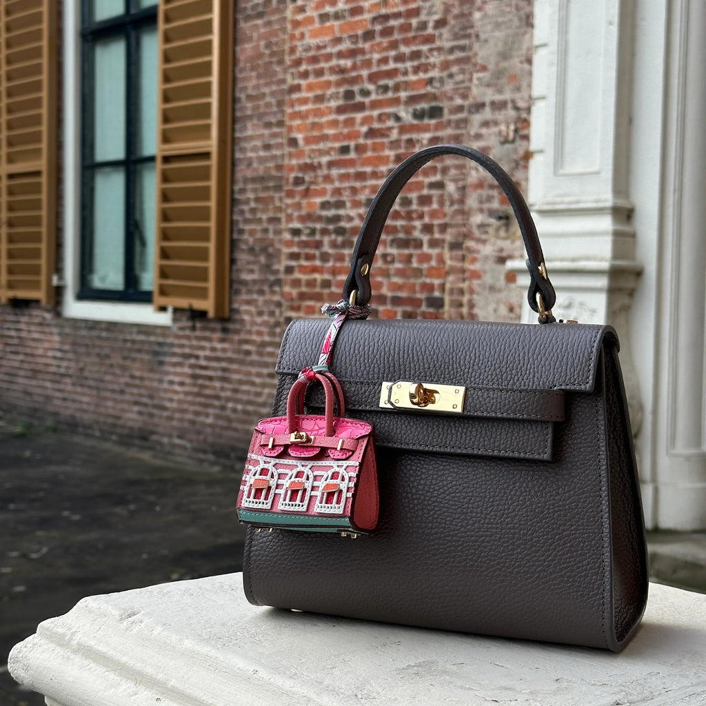 Gray handbag with a pink patterned bag charm on a stone ledge against a brick wall.