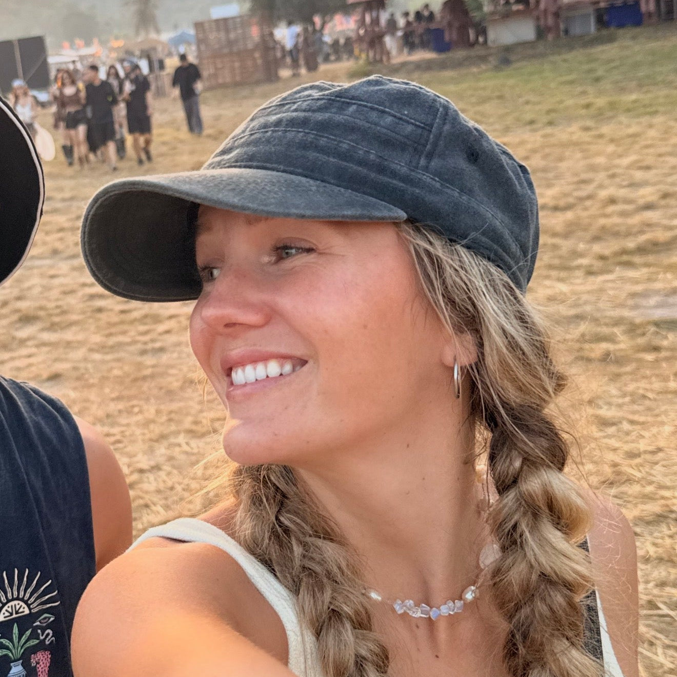 Woman wearing a black cap and white top with braided hair, standing outdoors with people and tents in the background.
