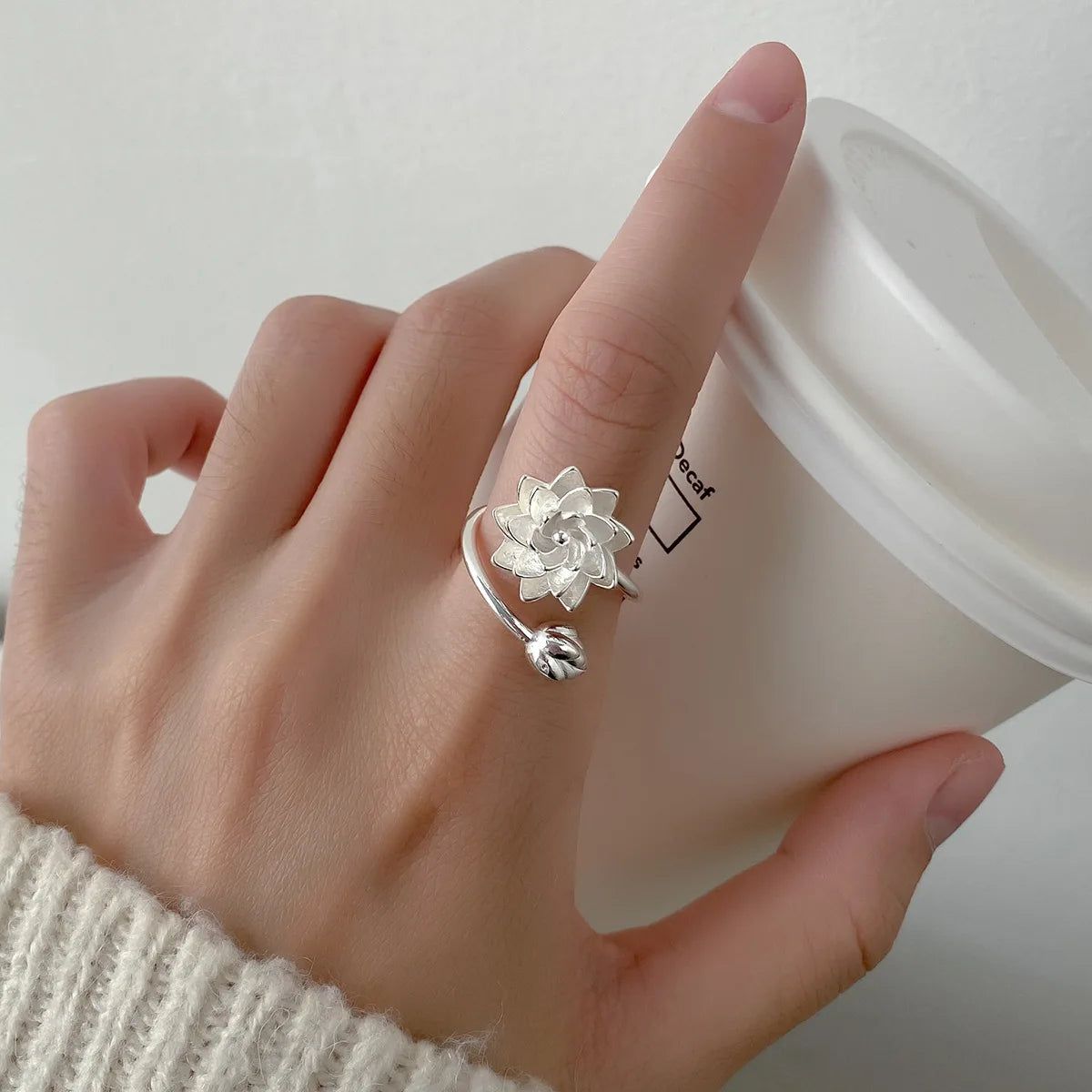 Hand wearing a silver lotus flower ring with a blurred coffee cup in the background