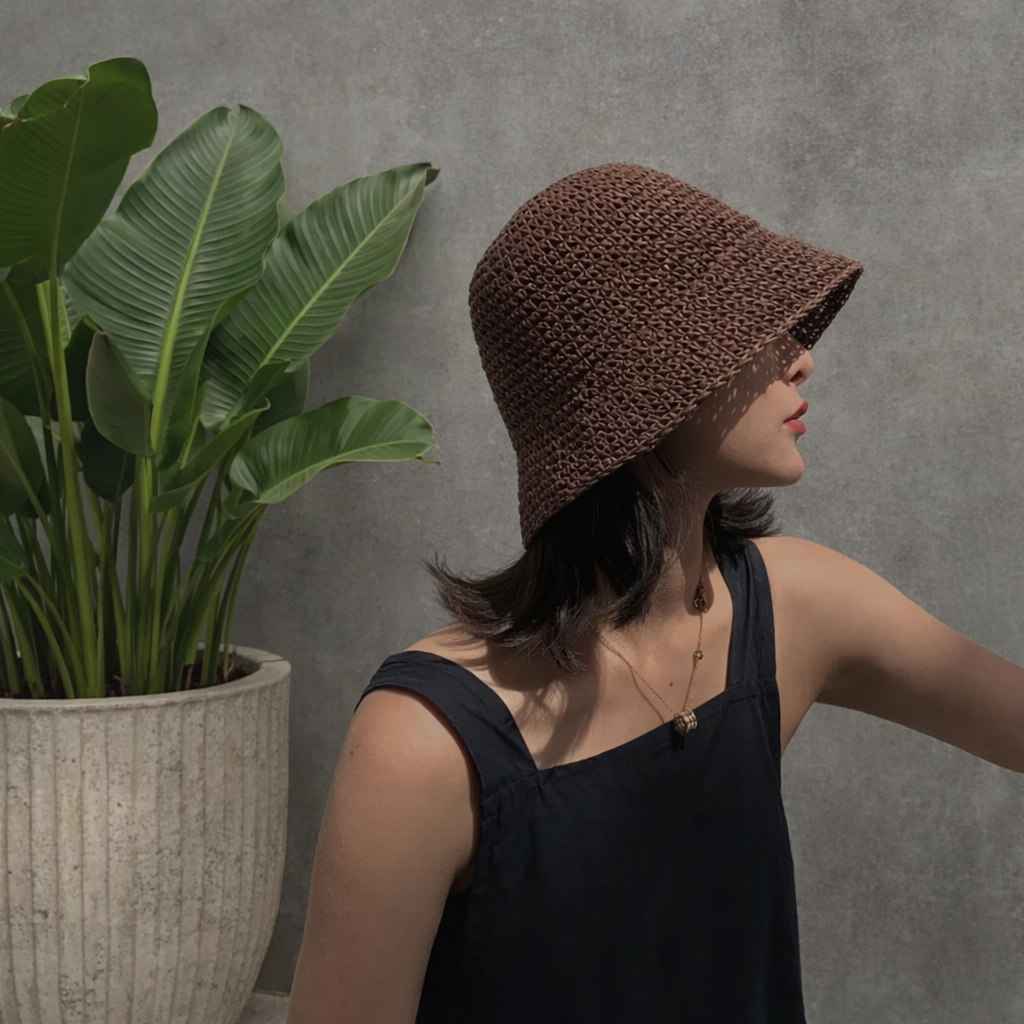 Woman wearing a brown knitted hat next to a potted plant against a gray wall