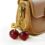 Gold cherry-shaped keychain attached to a beige handbag on a white background.
