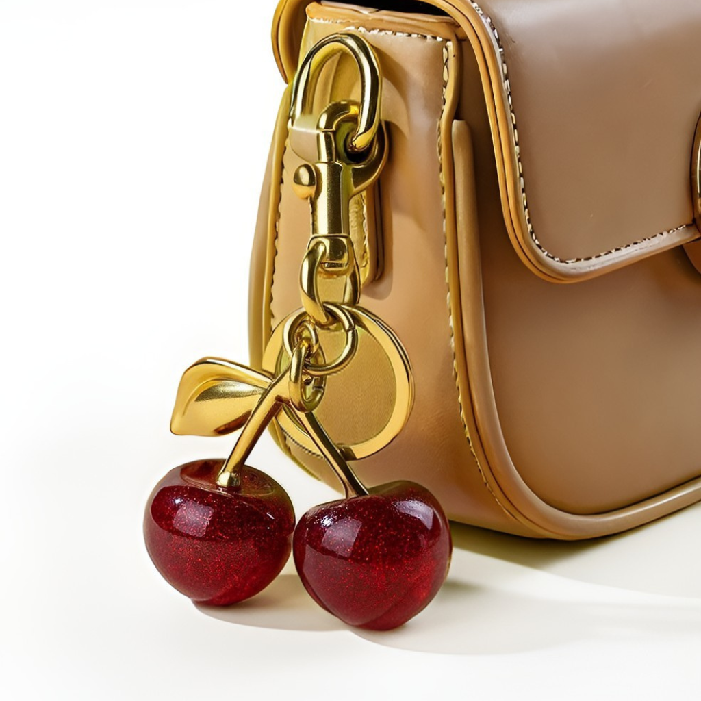Gold cherry-shaped keychain attached to a beige handbag on a white background.