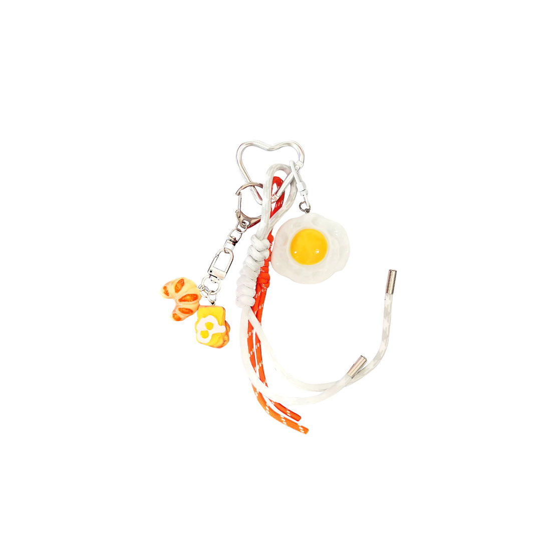 Novelty fried egg and croissant food charm on a keychain with white and orange-red cord detail.