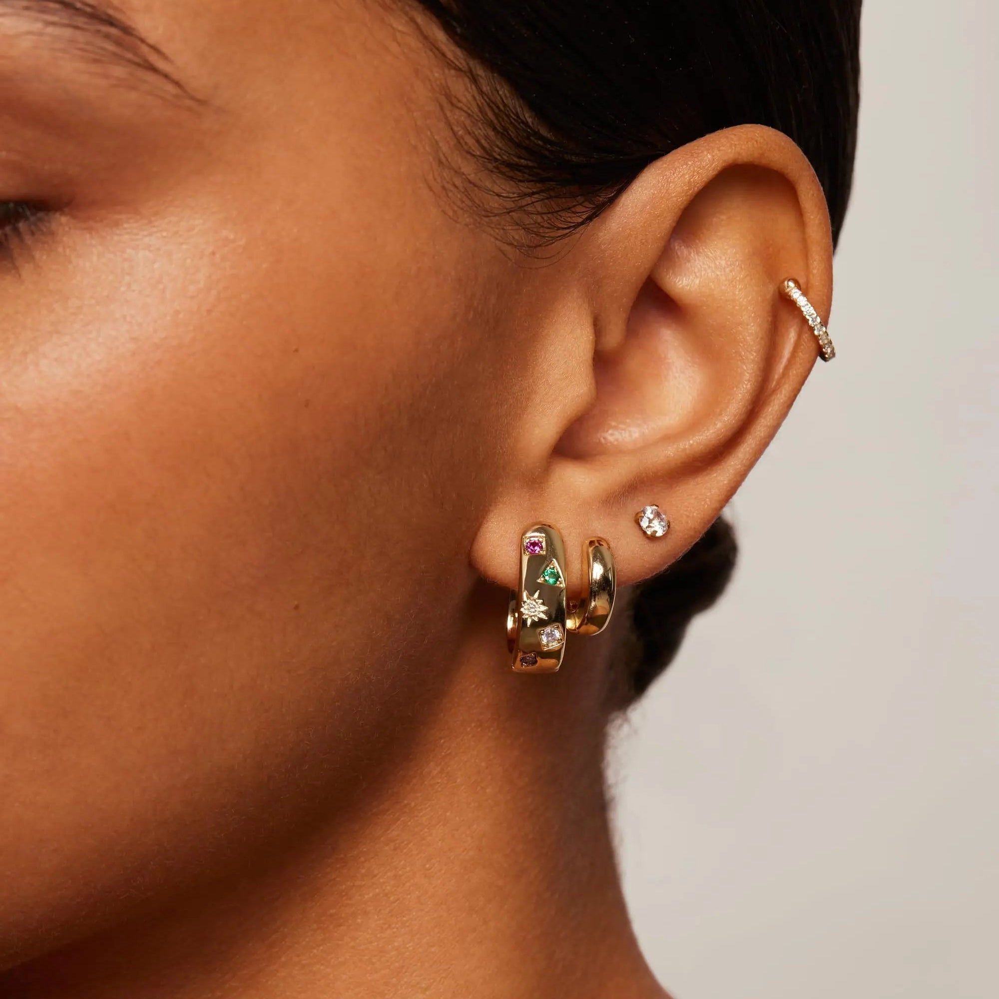 Close-up of a person wearing gold hoop earrings with green gemstones on a neutral background