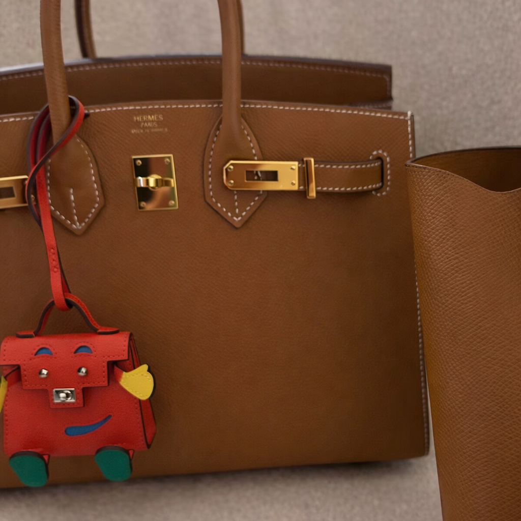 Brown leather handbag with gold hardware and a red dog-shaped charm on a beige surface.