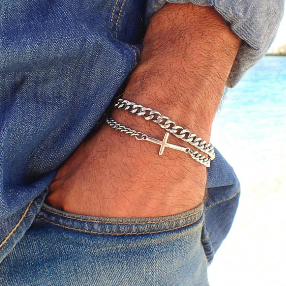 Silver chain bracelet with a cross on a person's wrist, wearing blue jeans, with a blurred background.