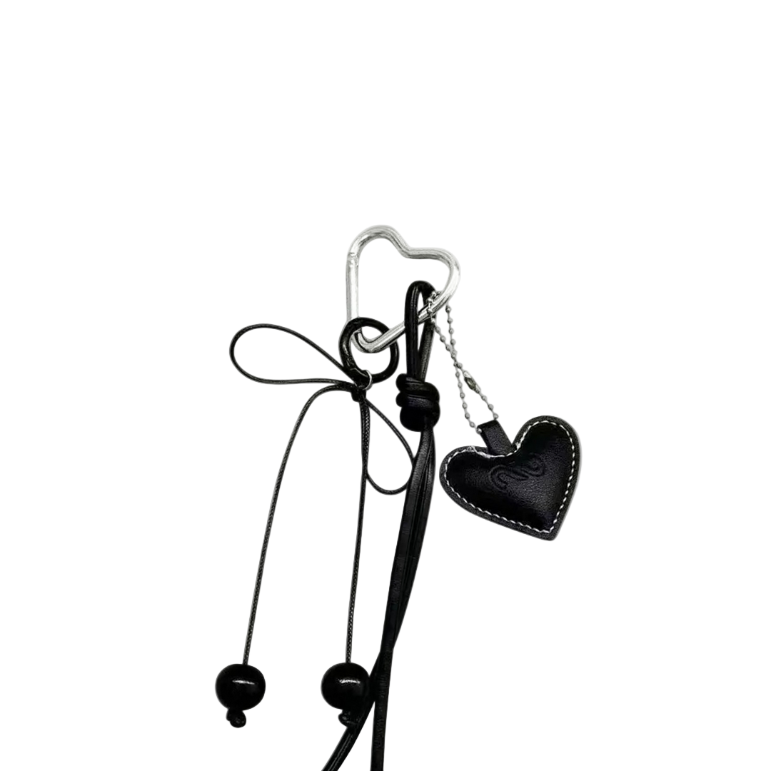 Black leather heart bag charm with white contrast stitching, hanging from a silver heart-shaped carabiner and black decorative cords.
