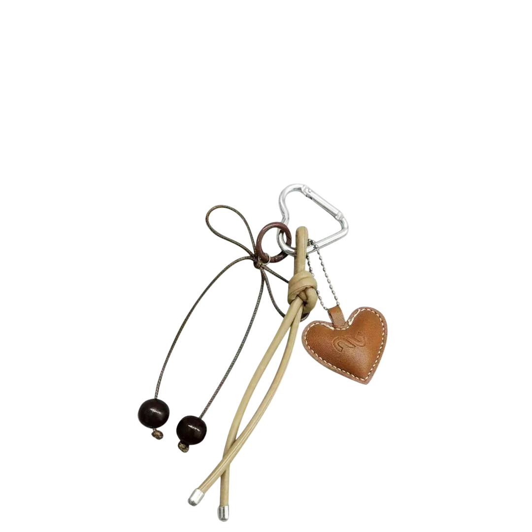 Brown leather heart bag charm with white contrast stitching, hanging from a silver heart-shaped carabiner and beige and brown decorative cords.