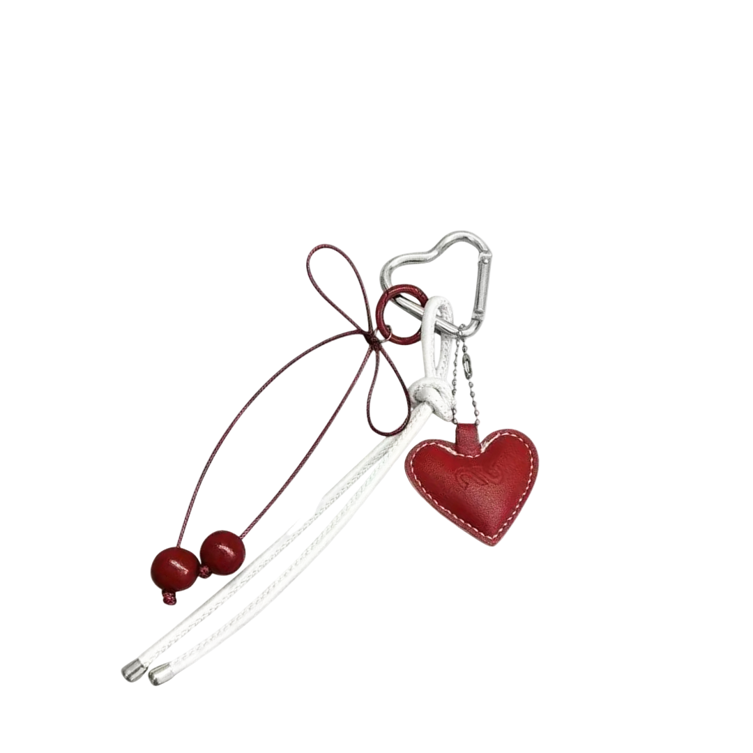 Red leather heart bag charm with white contrast stitching, hanging from a silver heart-shaped carabiner and red and white decorative cords.